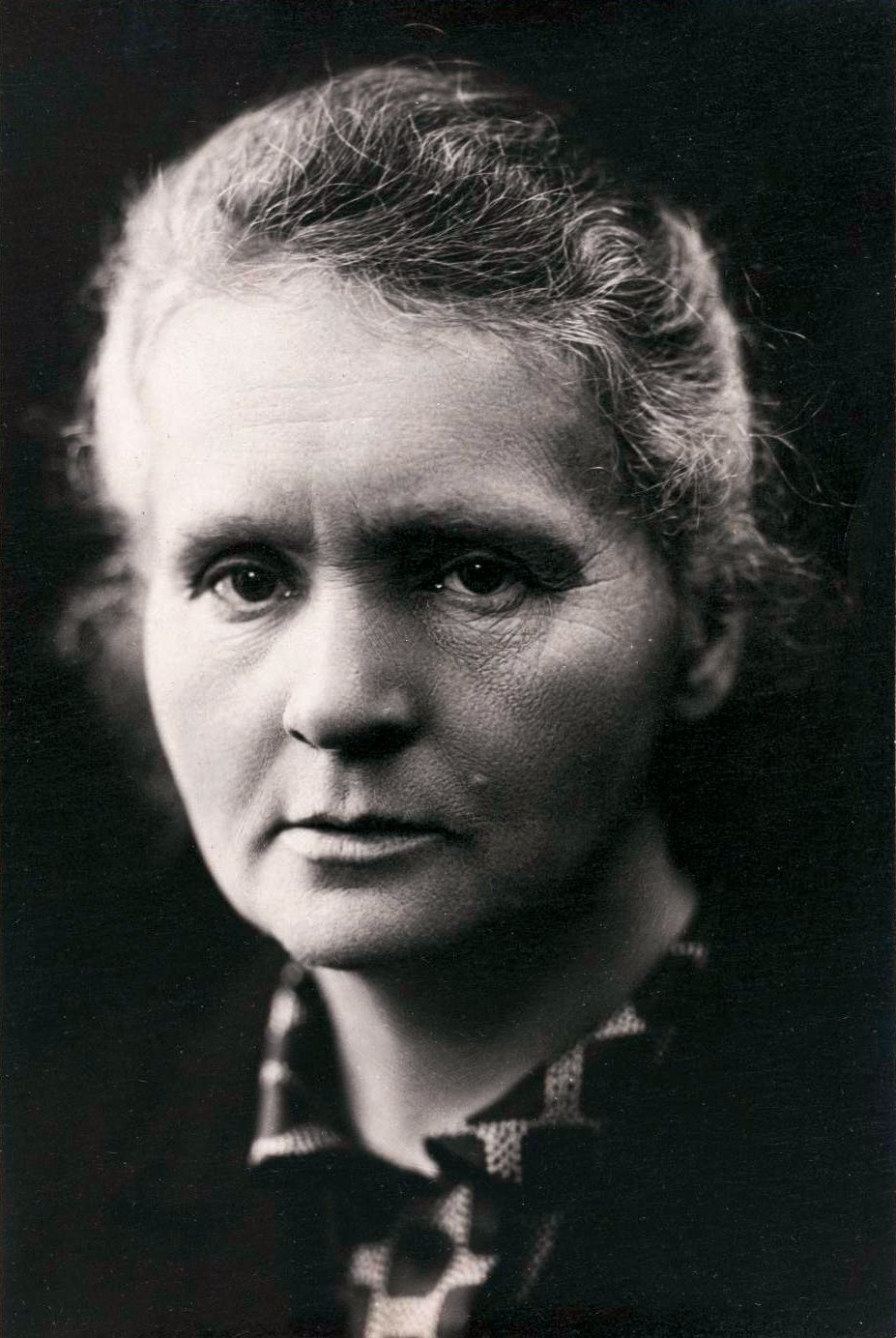 Retrato en blanco y negro de Marie Curie mirando a cámara, con el cabello recogido y una prenda oscura.